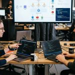 dos colegas de IT conversando frente a sus laptops y una pantalla