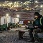 trabajador minero utiloizando una notebook en un campamento remoto frente a una operación minera iluminada durante la noche