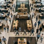 Vista aérea de una oficina moderna de planta abierta, con personas trabajando y desplazándose entre puestos de trabajo, en un entorno colaborativo y de desarrollo profesional