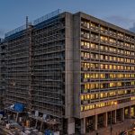 Edificio de oficinas en proceso de renovación, con andamios en una de sus fachadas mientras el resto de la estructura permanece operativo e iluminado