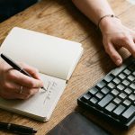 manos de hombre trabajando en simultáneo sobre un teclado y tomando notas en un cuaderno para ilustrar el riesgo de deskilling