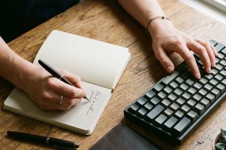 manos de hombre trabajando en simultáneo sobre un teclado y tomando notas en un cuaderno para ilustrar el riesgo de deskilling
