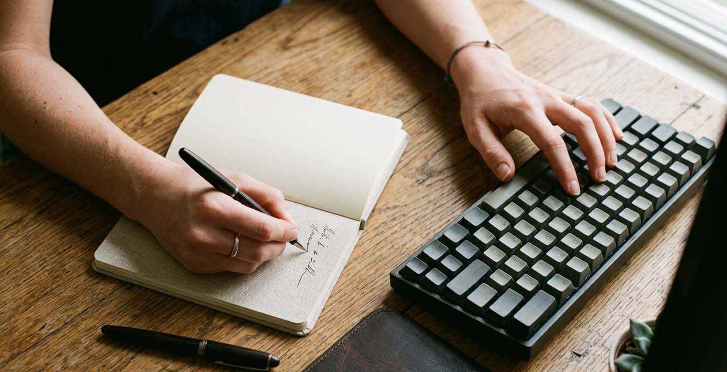 manos de hombre trabajando en simultáneo sobre un teclado y tomando notas en un cuaderno para ilustrar el riesgo de deskilling
