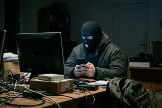 Persona con pasamontañas frente a una computadora sosteniendo un telefono representando una estafa vinculada a una oferta laboral falsa.