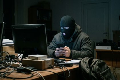 Persona con pasamontañas frente a una computadora sosteniendo un telefono representando una estafa vinculada a una oferta laboral falsa.