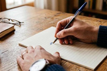 Primer plano de manos de un hombre escribiendo en un anotador, se lee un signo pesos entre signos de pregunta, para representar la duda al establecer una remuneracion pretendida